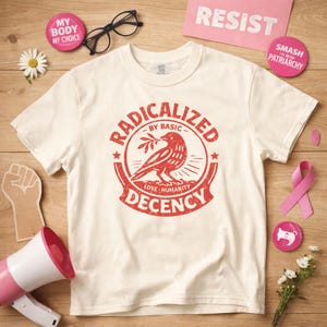 Puede incluir: Camiseta color crema con un gráfico rojo de un pájaro con una rama, rodeado por las palabras "RADICALIZED BY BASIC LOVE HUMANITY DECENCY." Otros elementos incluyen una cinta rosa, botones y la palabra "RESIST."