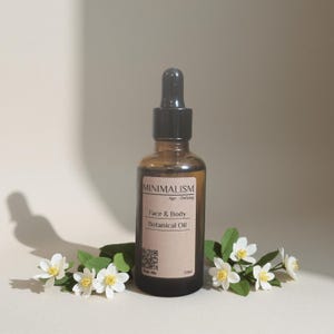 Könnte beinhalten: Eine braune Glasflasche mit "Face & Body Botanical Oil" und einer schwarzen Pipette, beschriftet mit "MINIMALISM Age-Defying". Die Flasche ist von weißen Blumen mit gelben Zentren und grünen Blättern umgeben. Die Flasche enthält 50 ml Öl.