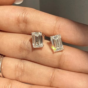 May include: A pair of emerald-cut diamond stud earrings. The earrings are set in a gold or platinum setting. The diamonds are clear and sparkling, reflecting light. The earrings are displayed on a hand with manicured nails.