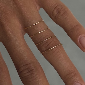 Puede incluir: Primer plano de una mano con cuatro anillos finos de oro. Los anillos están espaciados uniformemente en los dedos. La persona lleva una manga a rayas marrones y blancas.