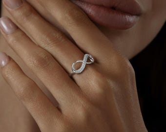Könnte beinhalten: Ein silberner Ring in Form eines Unendlichkeitszeichens. Der obere Bogen des Unendlichkeitszeichens ist mit kleinen, klaren Edelsteinen besetzt. Der Ring befindet sich an einem Finger einer Person, der Hintergrund ist unscharf.
