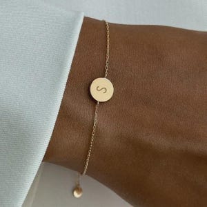Puede incluir: Una delicada pulsera dorada con un pequeño dije redondo con la letra "S" grabada. La pulsera tiene una fina cadena y un pequeño dije colgante. La joya se lleva en la muñeca.