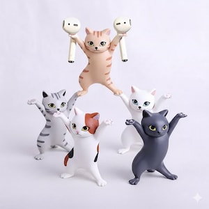 May include: A collection of cat-shaped figurines in various colors, including gray, white, and black, each with outstretched arms. One cat holds two white and beige earbud holders. The figurines have detailed features and are set against a white background.