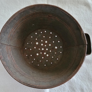 Op de afbeelding: Ooghoogte van een vintage, roestige metalen vergiet. Het ronde vergiet heeft een geperforeerde bodem en een klein handvat. Het metaal heeft een verweerde, verouderde uitstraling.