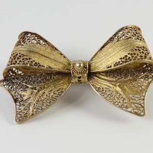 May include: A gold-colored filigree bow brooch. The brooch features an intricate openwork design, with a central knot and detailed bow loops. The metal has a slightly aged appearance, adding to its vintage charm. The brooch is a decorative accessory.