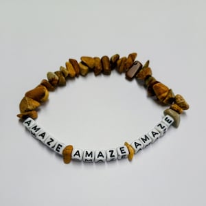 May include: A bracelet made of irregular, polished brown and tan stone chips interspersed with white square beads featuring the black printed text AMAZE. The beads are arranged in a repeating pattern with the word AMAZE appearing multiple times.