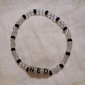 Op de afbeelding: Een kralenarmband met doorzichtige en zwarte kralen. De armband spelt de naam "NED" met witte letterkralen. De armband ligt op een wit oppervlak.