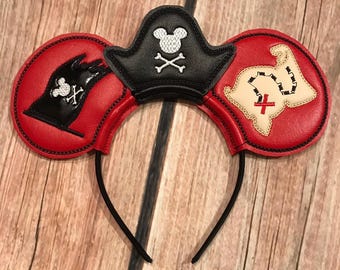 May include: Red and black pirate-themed mouse ears headband. The ears feature a black pirate hat with a skull and crossbones, a black glove with a skull and crossbones, and a map with a red cross. The headband is black.