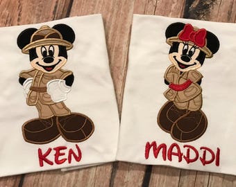 May include: Two white t-shirts with embroidered cartoon characters. One features a character in a safari outfit with the name "KEN" in red. The other has a character with a red bow and the name "MADDI" in red.