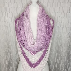 May include: A handmade crochet cowl scarf in shades of purple and white, displayed on a mannequin. The scarf features a gradient effect, transitioning from light purple to a deeper hue. The triangular shape and open weave design are visible.