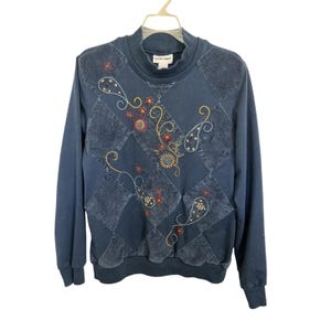 Pode incluir: Um moletom azul marinho com gola alta. A frente apresenta um design de patchwork com bordados paisley e padrões florais em tons de laranja, amarelo e azul. A parte superior de manga comprida está pendurada em um cabide branco.