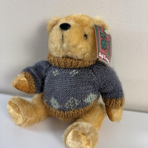 May include: A soft toy teddy bear with golden fur, dressed in a blue knitted sweater with a brown collar and cuffs. The sweater features a snowflake pattern. A tag reads "Soft Expressions".