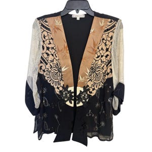 May include: A black, tan, and cream-colored open front jacket with floral and paisley patterns. The jacket has long sleeves with a sheer overlay and a decorative floral embellishment at the waist. The label reads "Spencer Alexis".