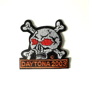 May include: Embroidered patch featuring a skull with red eye sockets and crossed bones. The skull is gray with black outlines, and the text "DAYTONA 2003" is displayed in orange on a black rectangular base.