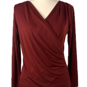 May include: A long-sleeved, burgundy wrap top with a V-neck. The fabric drapes across the chest and gathers at the side. The sleeves are long and fitted. The top is displayed on a mannequin.