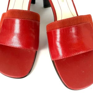 Puede incluir: Un par de sandalias de cuero rojo con punta cuadrada y una correa ancha en la parte superior. Las sandalias tienen un tacón de bloque y un detalle de gamuza en la correa. Los zapatos son de color rojo vibrante.