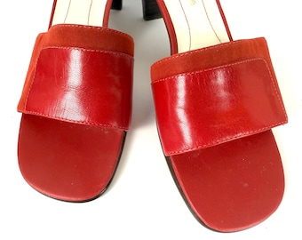 May include: A pair of red leather sandals with a square toe and a wide strap across the top. The sandals have a block heel and a suede accent on the strap. The shoes are a vibrant red color.