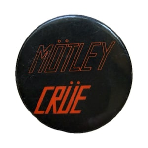May include: A round, black button with the band name "Mötley Crüe" in orange, outlined letters. The word "Mötley" is arched across the top, and "Crüe" is below it. The button has a glossy finish.