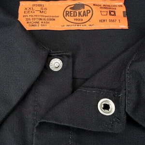May include: A close-up of a black work shirt collar with two silver snap closures. An orange tag displays the "RED KAP" logo and garment information, including fabric composition and care instructions. The shirt is made of 65% polyester and 35% cotton.