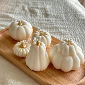 May include: Five off-white pumpkin-shaped candles with gold-colored stems and white wicks arranged on an oval wooden tray. The candles vary in size and shape, featuring ribbed textures that mimic the appearance of natural gourds.