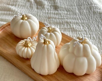 Puede incluir: Cinco velas de color blanco roto con forma de calabaza, con tallos dorados y mechas blancas, dispuestas sobre una bandeja ovalada de madera. Las velas varían en tamaño y forma, presentando texturas acanaladas que imitan el aspecto de las calabazas.