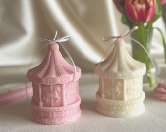 Puede incluir: Dos velas con forma de carrusel, una de color rosa claro y otra de color crema, cada una con figuras de caballos en relieve y un techo puntiagudo. Una cinta blanca fina está atada en la parte superior de la vela rosa y una cinta rosa en la crema.