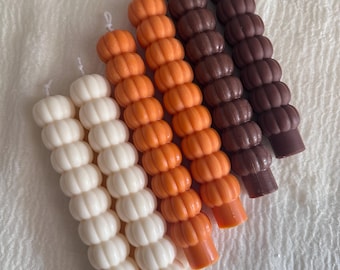 Puede incluir: Un conjunto de seis velas cilíndricas altas con una superficie texturizada y segmentada que recuerda a una pila de pequeñas esferas. Las velas están dispuestas en una paleta de colores degradados, con dos velas blancas, dos naranjas y dos de color marrón oscuro.