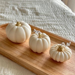 May include: Three off-white pumpkin-shaped candles of varying sizes, each featuring a metallic gold-painted stem and a white cotton wick. The candles are arranged in a row on an oval-shaped light-colored wooden tray with a visible natural grain.