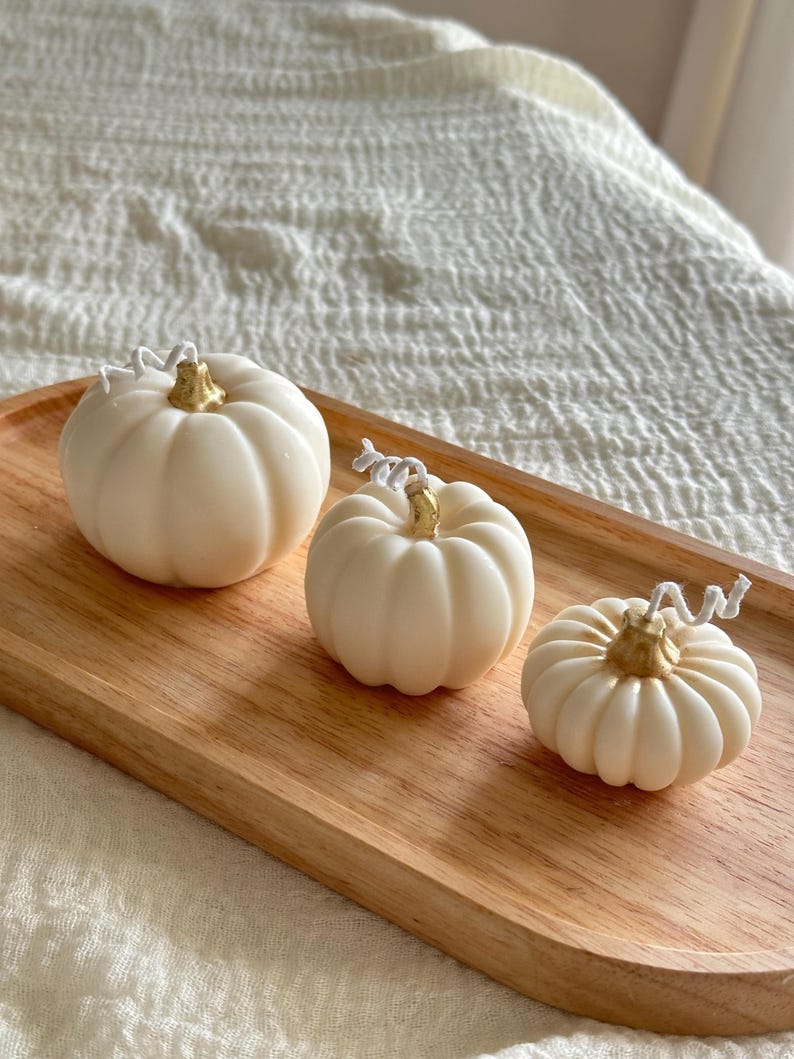 May include: Three off-white pumpkin-shaped candles of varying sizes, each featuring a metallic gold-painted stem and a white cotton wick. The candles are arranged in a row on an oval-shaped light-colored wooden tray with a visible natural grain.