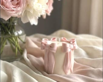 Puede incluir: Una vela cilíndrica de color blanco con lazos de color rosa claro en relieve adheridos a los lados. La vela tiene una única mecha centrada en la parte superior. Los lazos están esculpidos con largas cintas que caen por el lateral de la vela.