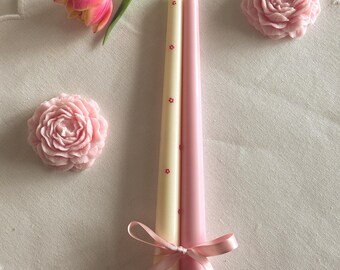 Puede incluir: Dos velas cónicas, una crema con detalles florales rojos, la otra rosa, atadas con una cinta rosa. Un tulipán rosa y dos decoraciones rosas en forma de rosa también están presentes. Las velas miden unos 25 cm de alto.