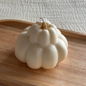 May include: A cream-colored, pumpkin-shaped candle with a ribbed, segmented surface. The top center features a small, metallic gold-painted stem detail surrounding a white cotton wick. The candle has a smooth, matte finish and a rounded base.