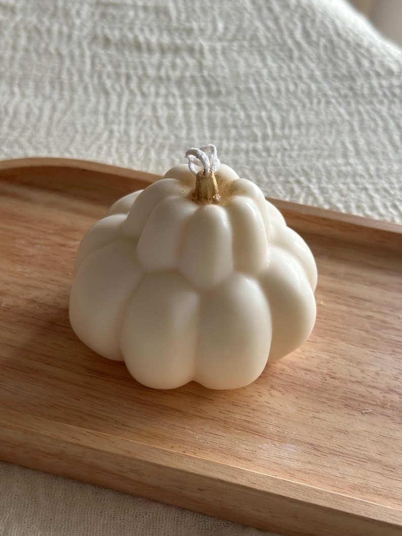 May include: A cream-colored, pumpkin-shaped candle with a ribbed, segmented surface. The top center features a small, metallic gold-painted stem detail surrounding a white cotton wick. The candle has a smooth, matte finish and a rounded base.