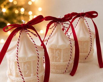 Puede incluir: Tres velas de color blanco roto con forma de carrusel y figuras de caballos moldeadas. Cada vela está rematada con un lazo de terciopelo rojo y un cordel trenzado rojo y blanco, creando un conjunto decorativo de adornos de cera.