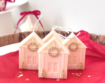Puede incluir: Tres velas de color rosa pálido con forma de casa y mechas blancas. Cada vela presenta una puerta de entrada moldeada, una corona dorada sobre la puerta y dos pequeños pinos dorados a ambos lados de la entrada, sobre papel de seda rojo.