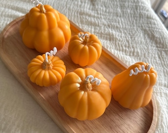 May include: Five orange pumpkin-shaped candles of varying sizes and shapes, each featuring a white cotton wick at the top. The candles are arranged on an oval-shaped light-colored wooden tray with a smooth, natural grain finish.