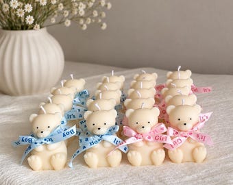 Puede incluir: Una colección de velas color crema con forma de oso dispuestas en filas. Cada vela tiene una pequeña mecha y está atada con una cinta impresa con 'It's a Boy' en azul o 'It's a Girl' en rosa, con un jarrón blanco de flores detrás.