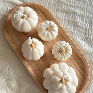 May include: Five off-white pumpkin-shaped candles with gold-painted centers and white wicks, arranged on an oval wooden tray. The candles vary in size and ribbed texture, featuring distinct segments and a smooth wax finish on a light wood base.