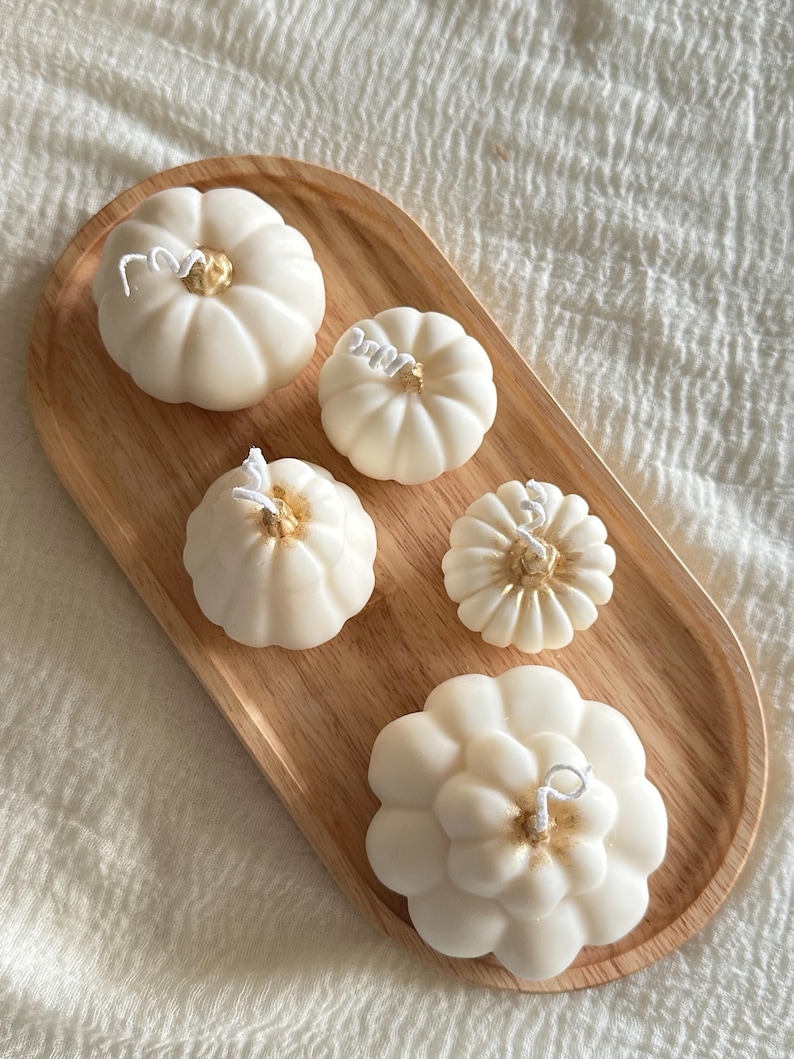 May include: Five off-white pumpkin-shaped candles with gold-painted centers and white wicks, arranged on an oval wooden tray. The candles vary in size and ribbed texture, featuring distinct segments and a smooth wax finish on a light wood base.