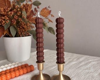 Puede incluir: Dos velas de pilar altas de color marrón oscuro con textura de burbujas y mechas blancas, cada una colocada en un portavelas de metal dorado mate liso con una base circular ancha. Las velas presentan un diseño segmentado y redondeado.