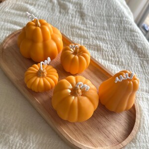 May include: Five orange pumpkin-shaped candles of varying sizes and shapes, each featuring a white cotton wick at the top. The candles are arranged on an oval-shaped light-colored wooden tray with a smooth, natural grain finish.