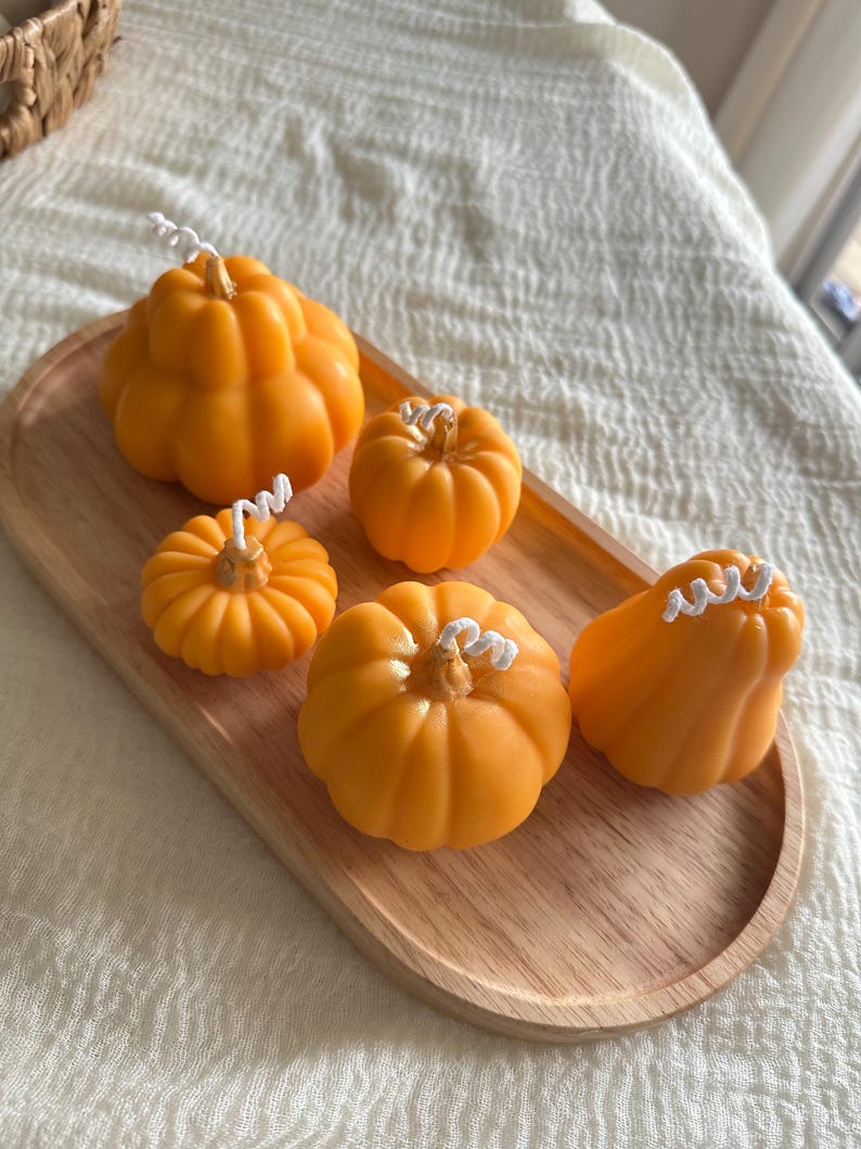 May include: Five orange pumpkin-shaped candles of varying sizes and shapes, each featuring a white cotton wick at the top. The candles are arranged on an oval-shaped light-colored wooden tray with a smooth, natural grain finish.