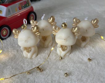 以下が含まれることがあります： 金色の角と装飾的なアクセントが付いた、4つのアイボリー色のトナカイのキャンドル。各キャンドルには笑顔の顔と小さな金のベルが付いています。赤いおもちゃの車が背景に見え、休日のテーマを示唆しています。