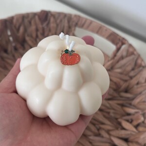 May include: A cream-colored, rounded, multi-lobed candle with a white wick. A small, orange, metal pumpkin-shaped charm with a green stem detail is attached to the top of the candle near the wick, resting against the smooth, wax surface.
