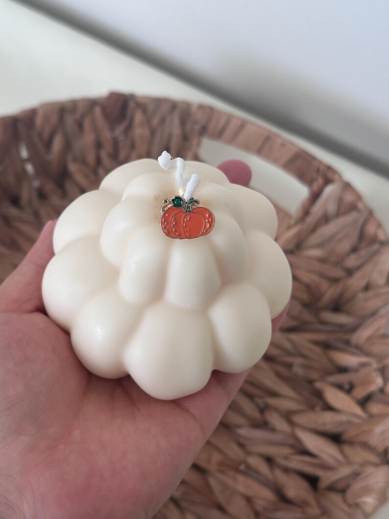 May include: A cream-colored, rounded, multi-lobed candle with a white wick. A small, orange, metal pumpkin-shaped charm with a green stem detail is attached to the top of the candle near the wick, resting against the smooth, wax surface.