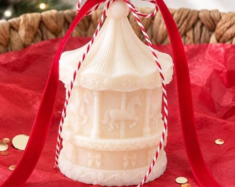 Puede incluir: Una vela de color crema con forma de carrusel que presenta figuras de caballos en relieve y detalles de lazos. La vela está rematada con una cinta de terciopelo rojo y un lazo de cordel a rayas rojas y blancas, con una mecha blanca central.