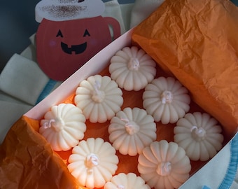 Puede incluir: Una caja rectangular que contiene diez velas blancas con forma de calabaza sobre papel de seda naranja. Junto a la caja hay una tarjeta decorativa con una taza de calabaza rosa con nata montada y una pajita marrón sobre tela blanca.