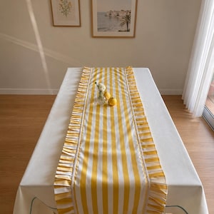 Puede incluir: Un camino de mesa rectangular con rayas verticales amarillas y blancas y bordes con volantes en ambos lados. El camino está centrado sobre un mantel blanco, con un pequeño florero de cristal con una flor blanca y dos limones amarillos.
