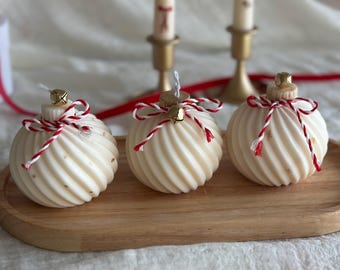 Puede incluir: Tres velas con forma de adorno navideño de color crema, con un patrón en espiral, lazos de cuerda rojos y blancos y pequeñas campanas doradas. Las velas están sobre una bandeja de madera. Dos velas están en el fondo, junto con cinta roja y candelabros dorados.