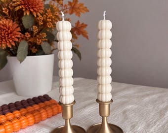 Puede incluir: Dos velas cilíndricas, altas y de color blanco roto, con una textura apilada de formas redondeadas similares a cuentas. Cada vela se sostiene verticalmente en un candelabro de metal dorado mate con una base circular ancha y un tallo fino.
